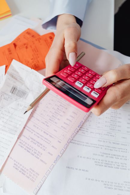 A close-up of a person's hands holding a pink calculator over a cluttered desk with scattered receipts, bills, and a pen, indicating financial and tax-related paperwork. The person is dressed in a light grey suit, and the background includes a white surface and some orange-colored paper. The scene suggests organized review and calculations associated with domestic or commercial cleaning services, aligning with Deep Cleaning Ilford's focus on surface cleaning, sanitisation, and hygiene maintenance within residential or commercial environments.