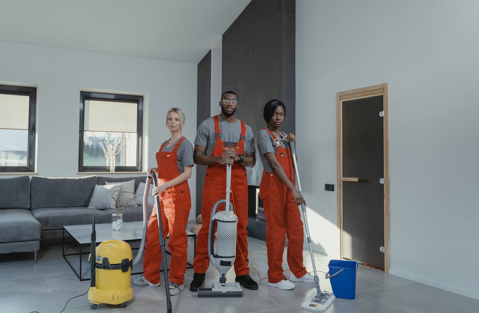 Three professional cleaners from Deep Cleaning Ilford are performing surface cleaning and deep cleaning in a modern living room. The team, composed of two women and one man, is dressed in grey uniforms with bright orange aprons, equipped with cleaning tools such as vacuum cleaners, mops, and buckets. They are standing in a well-lit, tidy living space with large windows allowing natural light to illuminate the grey tiled floor and a dark accent wall. The room features a contemporary grey sofa with cushions, a glass coffee table, and a wooden-framed chalkboard on one wall, all maintained in immaculate condition, reflecting thorough sanitisation and cleaning. Deep Cleaning Ilford's team is actively engaged in cleaning surfaces, ensuring hygiene and freshness in the environment, consistent with their domestic and commercial cleaning services for tenants in Seven Kings, Ilford.