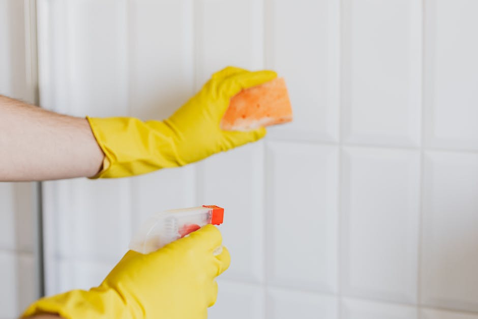 Close-up of a person wearing yellow rubber gloves performing surface cleaning on white ceramic tiled wall in a kitchen or bathroom. The individual is holding an orange sponge in one hand and a spray bottle in the other, applying a cleaning solution to the tiles. The tiles are clean and shiny, with visible grout lines, and the lighting highlights the pristine condition of the surface. This scene illustrates professional deep cleaning and sanitisation processes carried out by Deep Cleaning Ilford, aligned with services for Seven Kings tenants, ensuring hygiene and maintenance of domestic or commercial spaces.