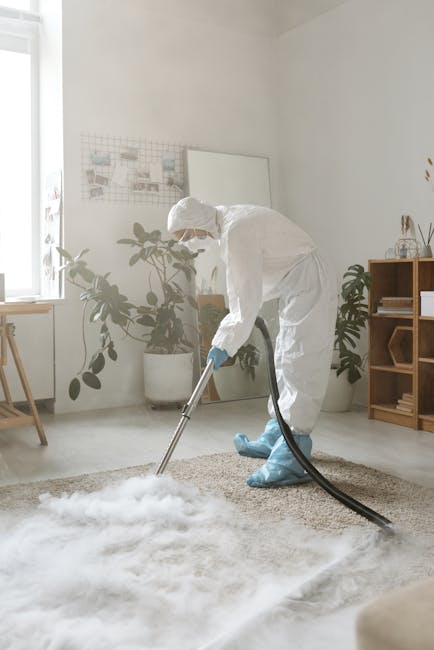 A professional cleaner dressed in full protective clothing, including a white suit, gloves, mask, and shoe covers, is performing deep carpet cleaning in a bright, modern living room. The cleaner uses a specialized upright vacuum or extractor machine, with a hose attachment directed at a plush, beige carpet that appears to be wet or being cleaned. The room features a large window allowing natural light, with green potted plants and minimalistic wooden furniture, including a shelf with small decorative items and a side table. The white walls and light-colored flooring contribute to a clean, fresh appearance, emphasizing hygiene and maintenance standards typical of domestic cleaning services offered by Deep Cleaning Ilford near Valentines Park Ilford. The scene reflects thorough surface cleaning and sanitization processes aimed at deep cleaning carpets and enhancing indoor cleanliness.