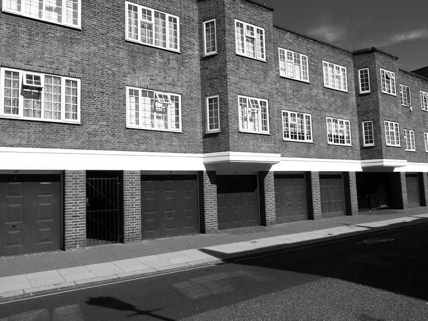 Black and white photograph of a multi-storey residential building with brick exterior walls and multiple white-framed windows. The ground level features several closed garage doors made of metal, set into brick columns that separate each parking space. A narrow sidewalk runs parallel to the building, with a shadow cast across the street in the foreground. The overall scene is well-lit with sharp contrast, highlighting the clean and orderly appearance of the building façade, indicative of regular exterior surface cleaning and maintenance by Deep Cleaning Ilford as part of their domestic cleaning services for flats on Cranbrook Road Ilford.