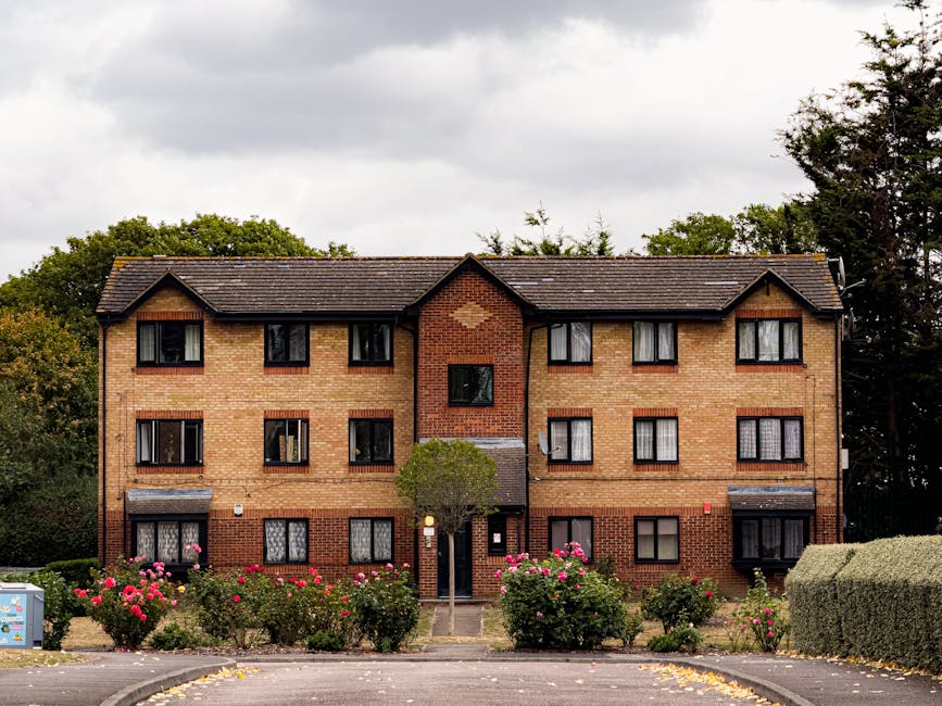 An external view of a three-story brick residential building with multiple windows, some featuring dark window frames and small awnings. The building is surrounded by well-maintained pink rose bushes and green shrubbery, with a small central pathway leading to the entrance door. The sky overhead is overcast with grey clouds, and the scene is lit evenly, highlighting the cleanliness of the exterior. This facade showcases the importance of regular surface cleaning and maintenance for residential properties, as provided by Deep Cleaning Ilford, as seen on the deepcleaningilford.co.uk website.